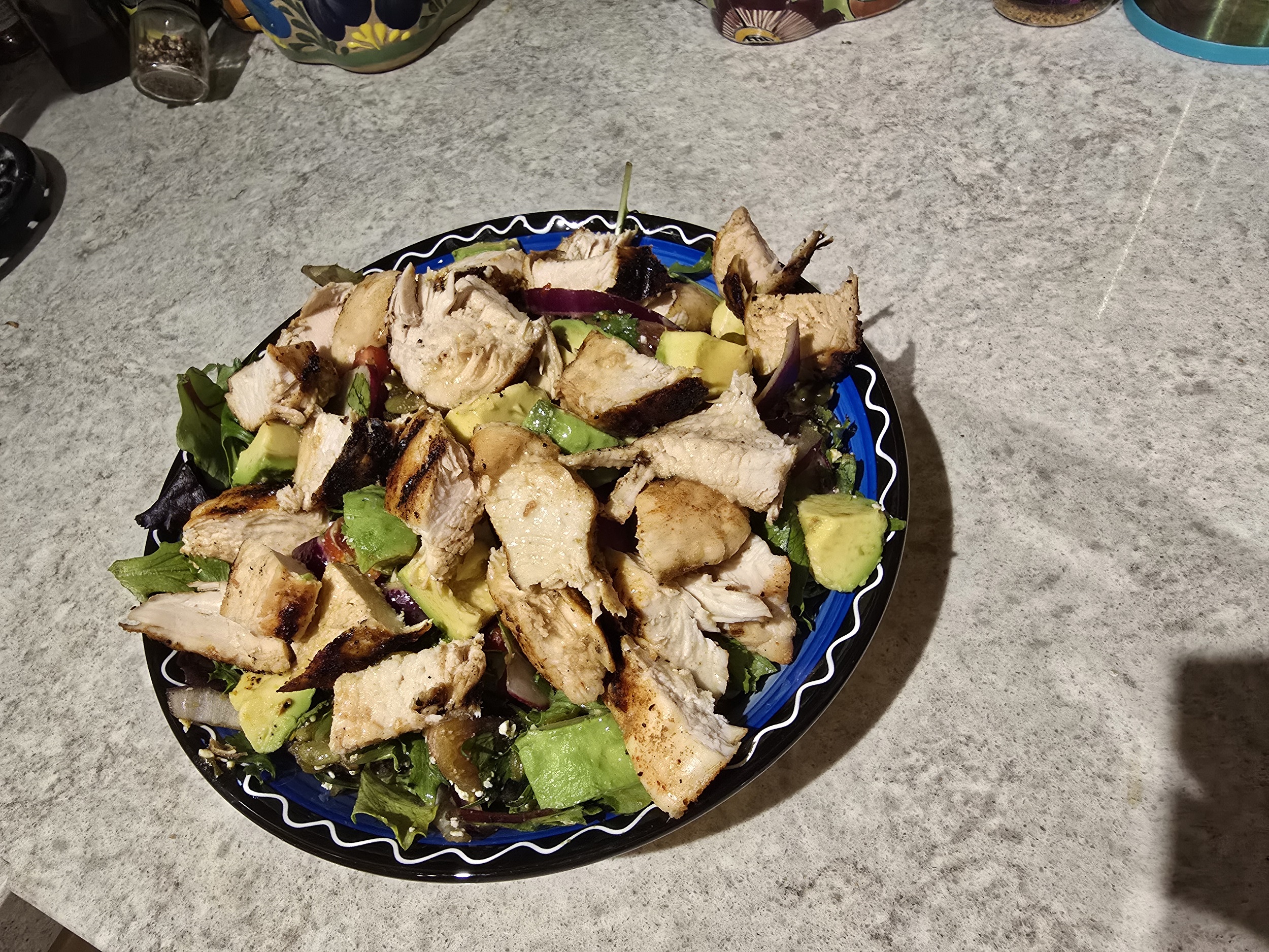 Overhead view of a homemade grilled chicken salad on a blue and black Talavera plate, served on a granite countertop. The salad features grilled chicken, avocado, red onion, mixed greens, and Cotija cheese.