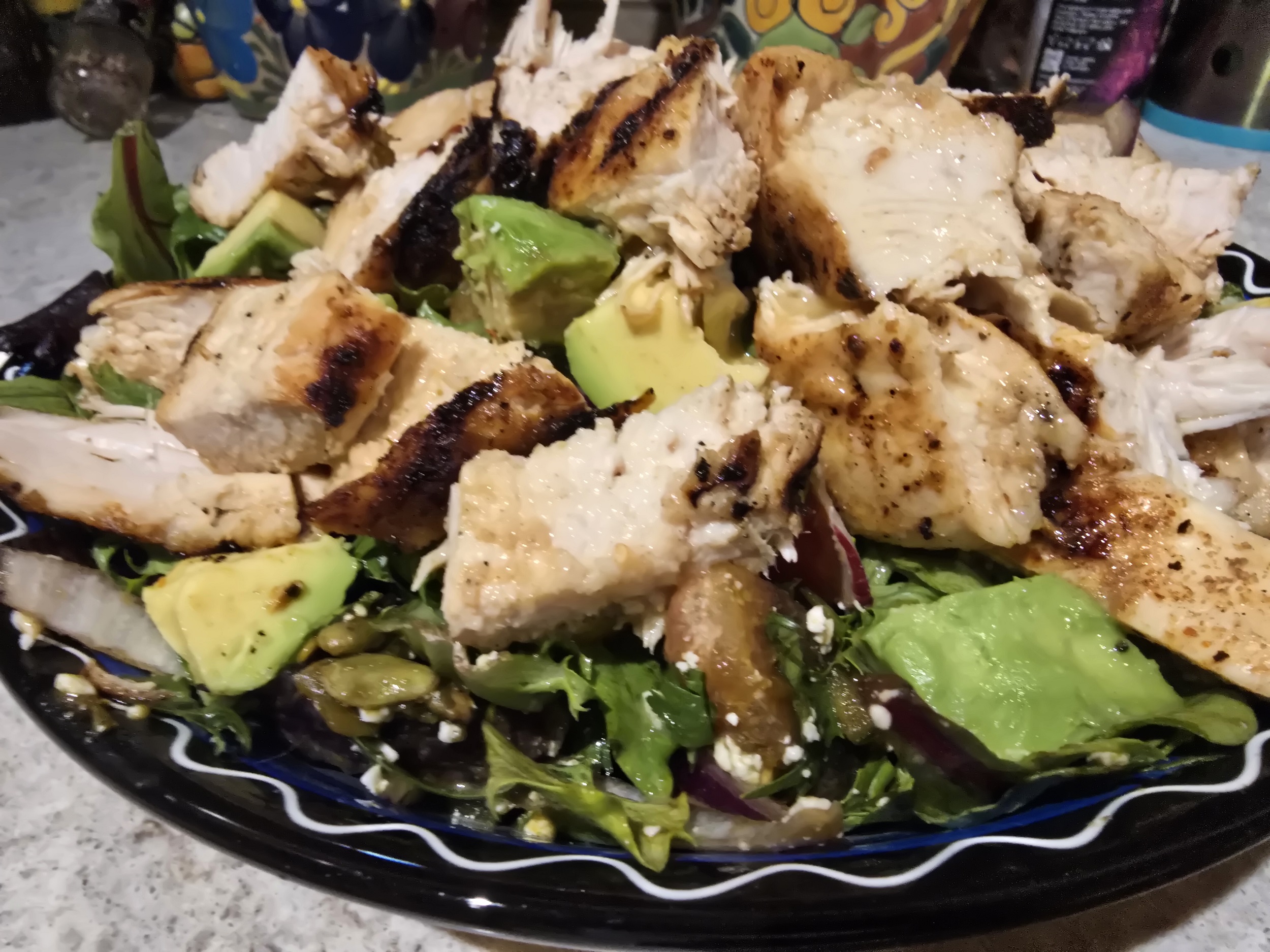 Close-up of a colorful Tucson-style salad piled high on a hand-painted blue and black Talavera plate, featuring chunks of grill-marked chicken, fresh avocado, mixed greens, red onion, pepitas, and crumbled Cotija cheese.