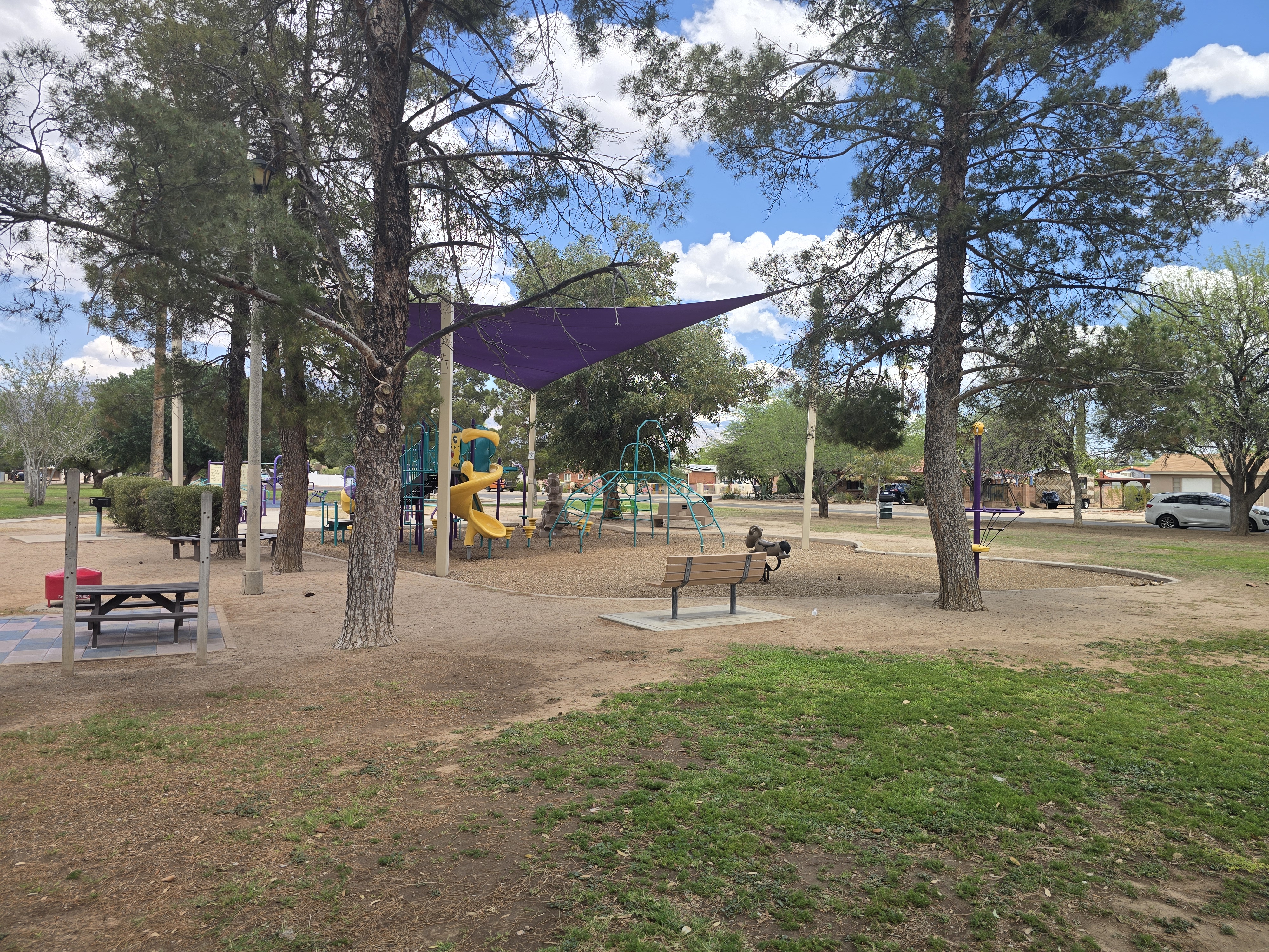 La Madera Park playground under a purple sail canopy, with a yellow slide, teal climbing dome, benches, and tall pines.