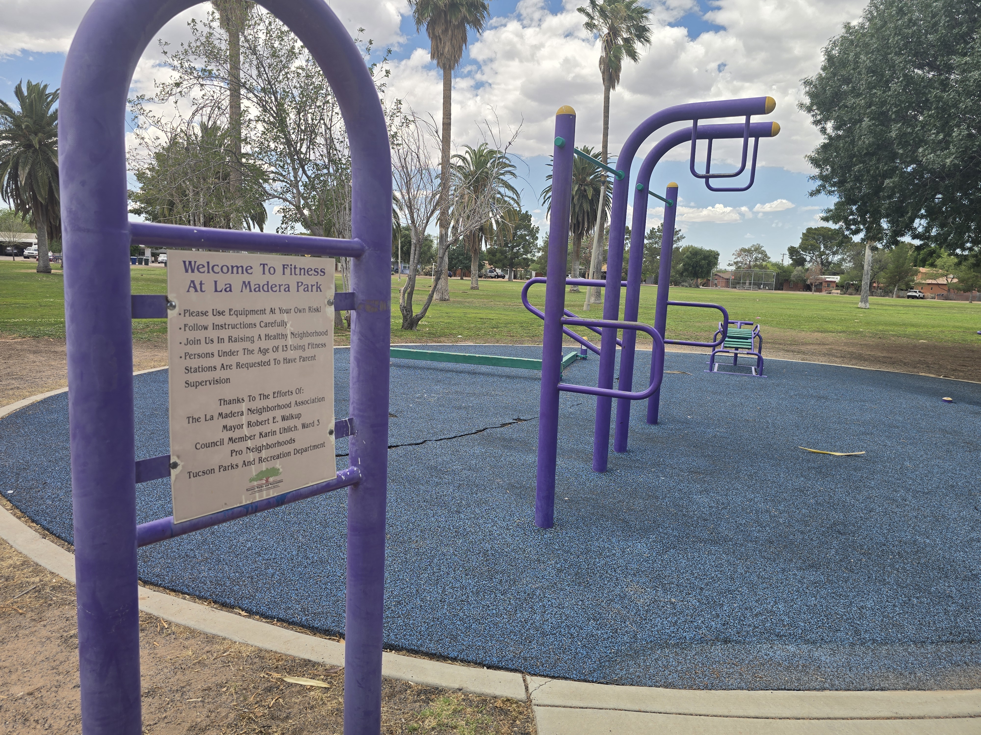 "Welcome to Fitness at La Madera Park" sign beside purple outdoor exercise equipment, with palm trees and a green lawn behind.