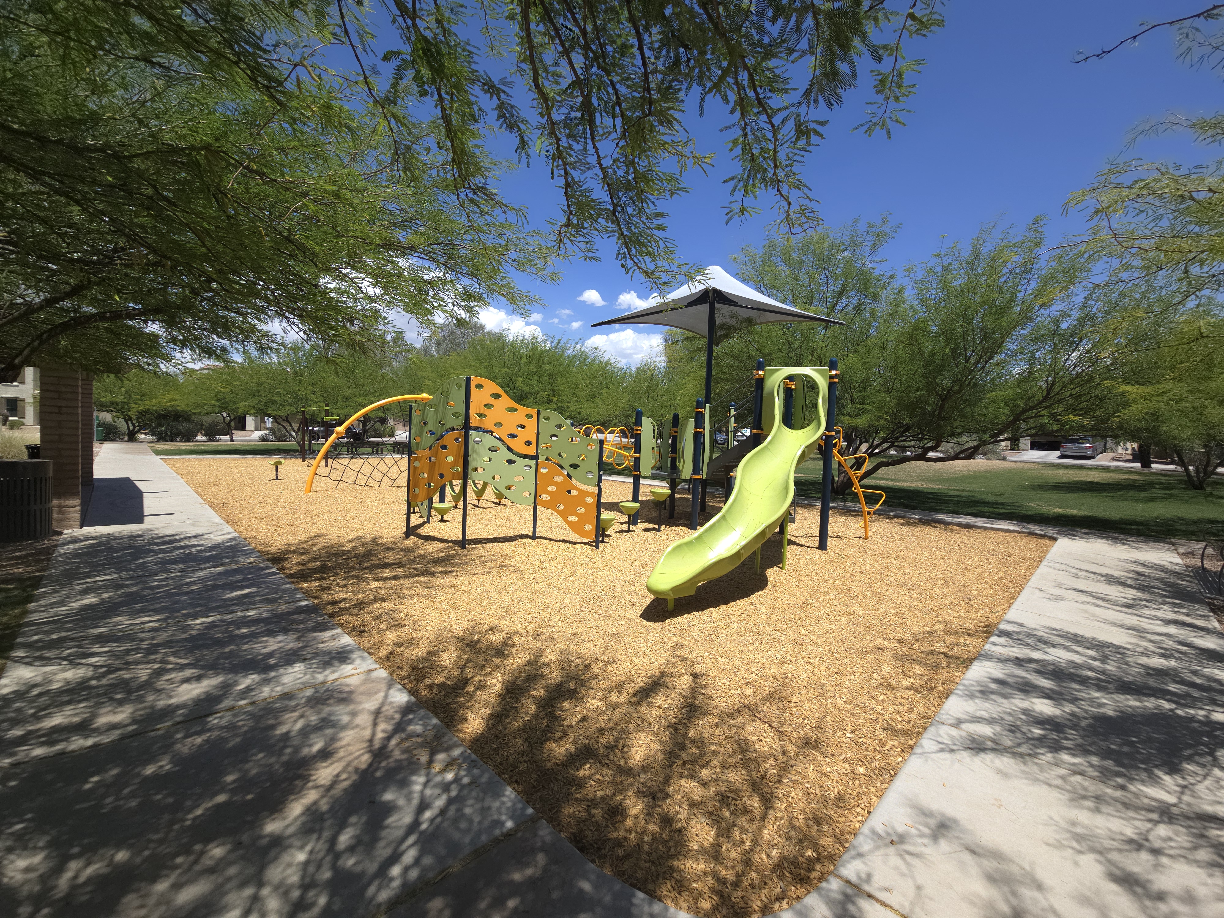 Modern playground at La Estancia de Tucson featuring a green slide, climbing wall, and shade sail surrounded by mesquite trees