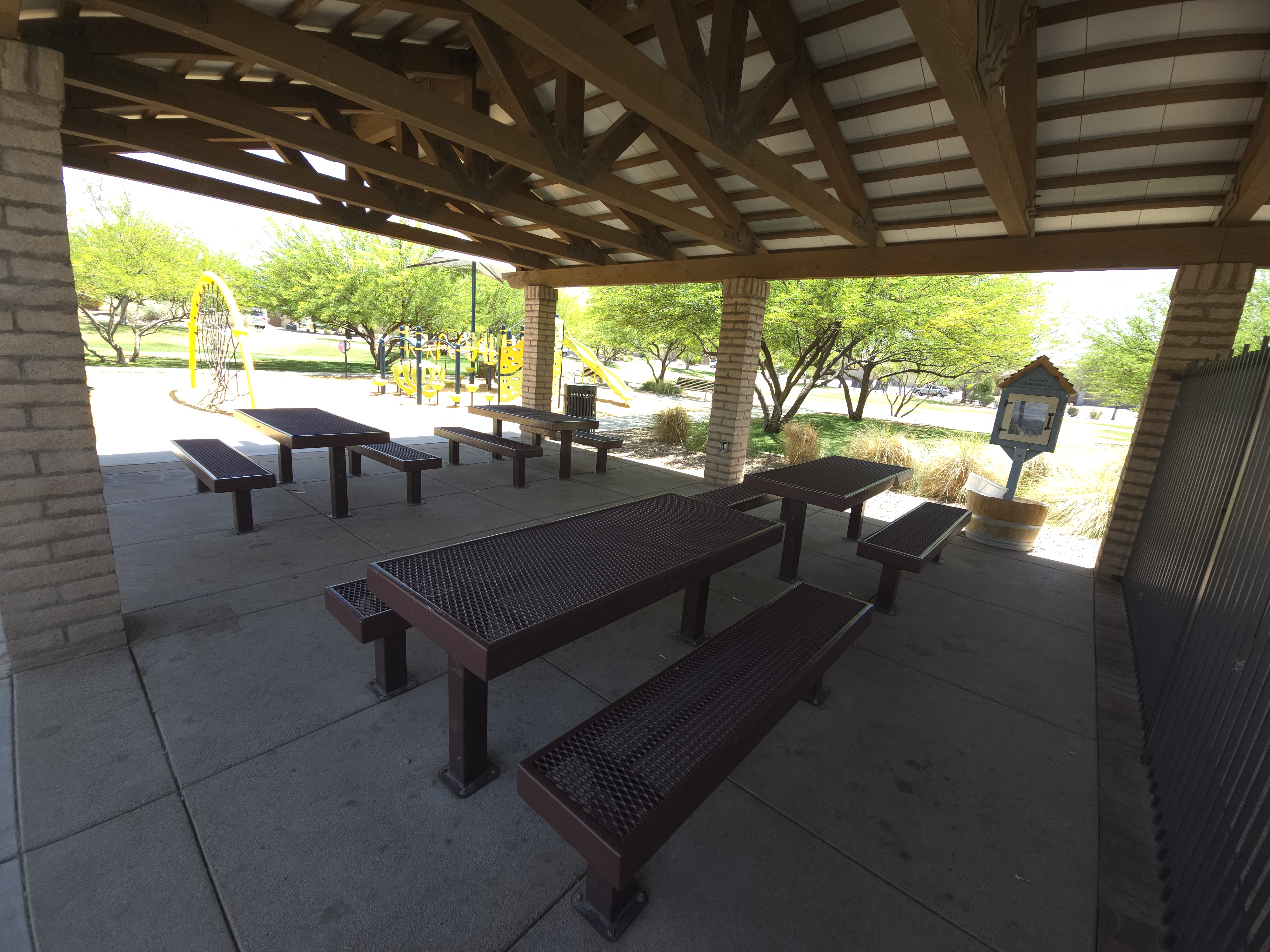 Covered picnic ramada with metal tables overlooking a yellow and blue playground and Little Free Library at a La Estancia community park