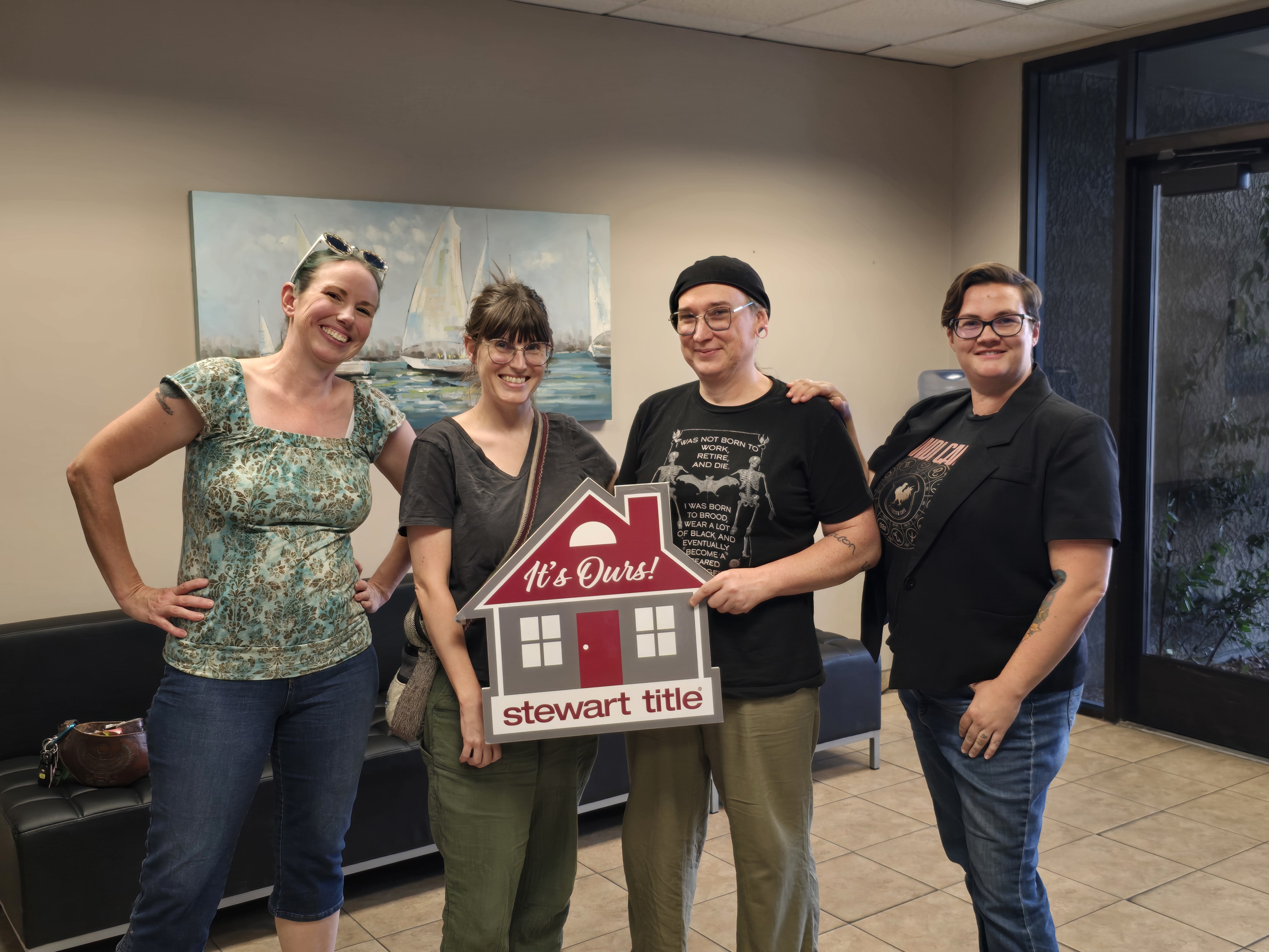 Four smiling people in a title company office, two holding a red house-shaped "It's Ours!" Stewart Title sign to celebrate a home purchase.