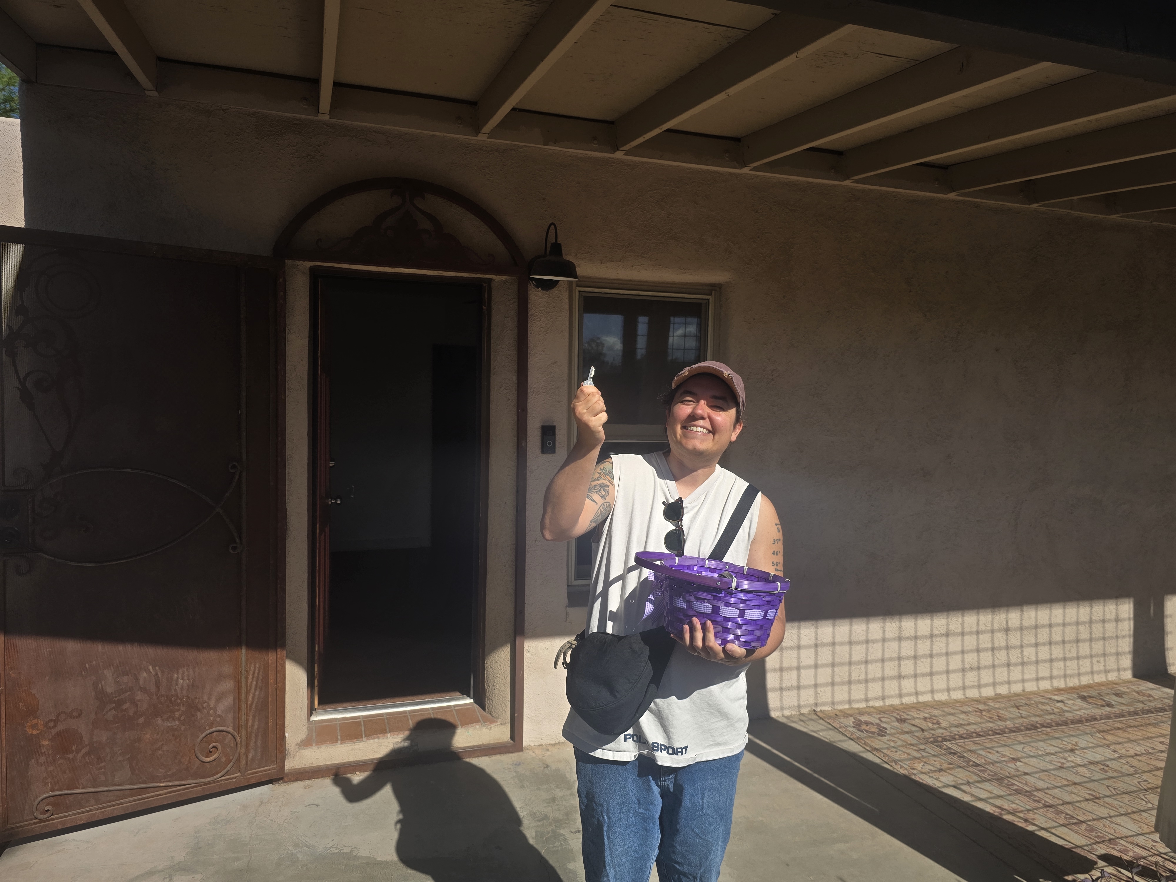 Person on a covered front porch holding up a house key and a purple woven basket, smiling beside an open front door.