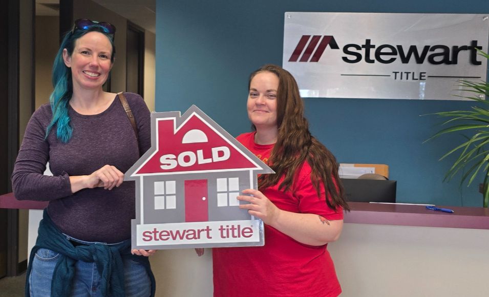 Two smiling women in the Stewart Title office lobby holding a red house-shaped "Sold" sign on closing day.
