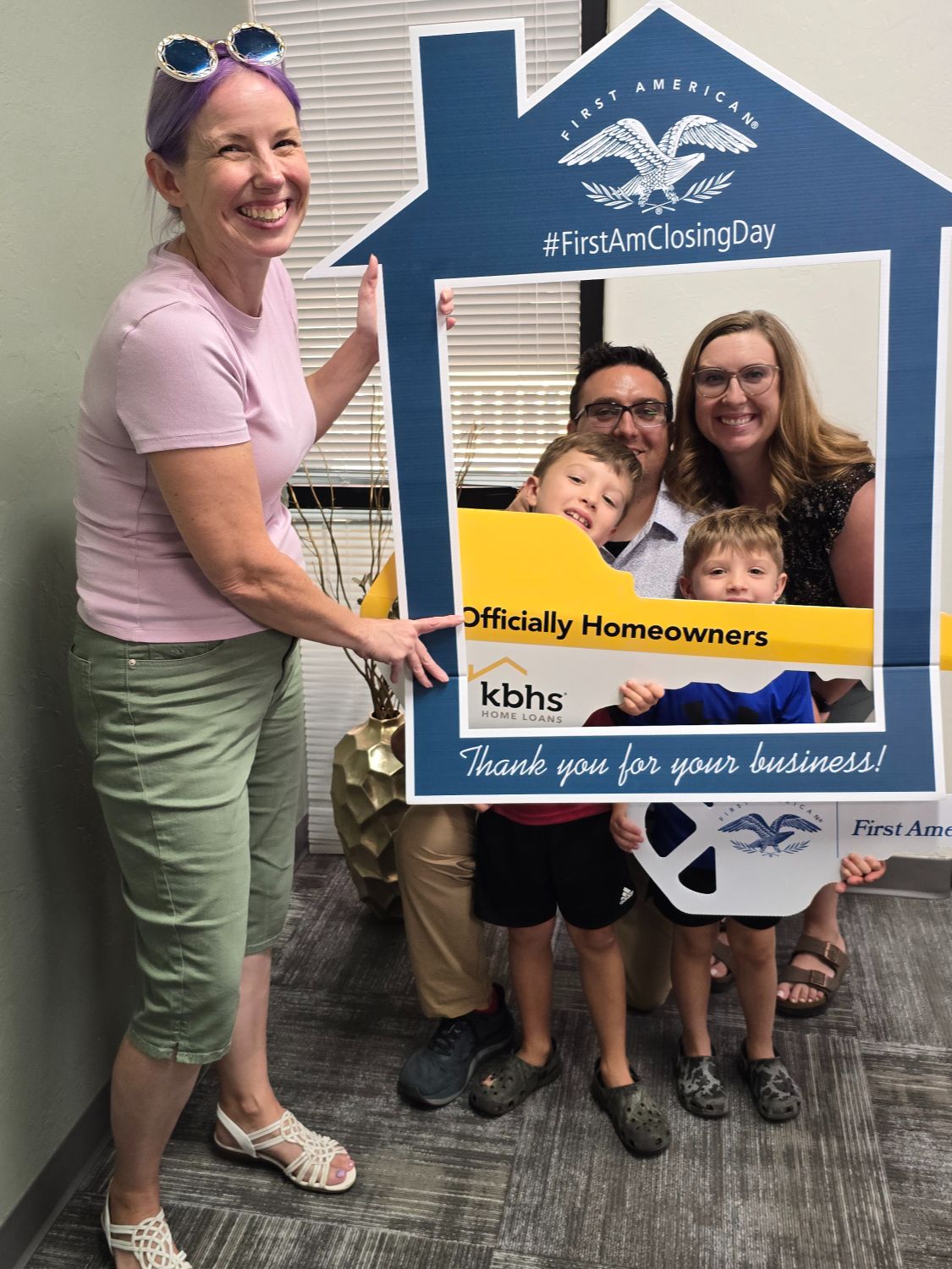 Woman posing beside a family of four inside a large "#FirstAmClosingDay – Officially Homeowners" photo frame at a title company closing.