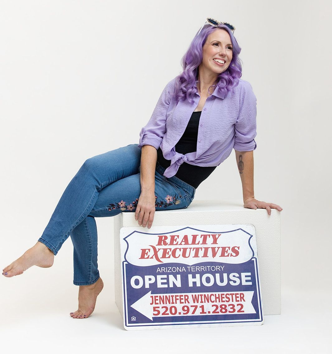 Jennifer Winchester sitting on a white block beside a Realty Executives Arizona Territory open house sign that reads "Jennifer Winchester, 520-971-2832." She has long wavy purple hair, a lavender button-down shirt, and jeans, and is smiling and looking off to the side.