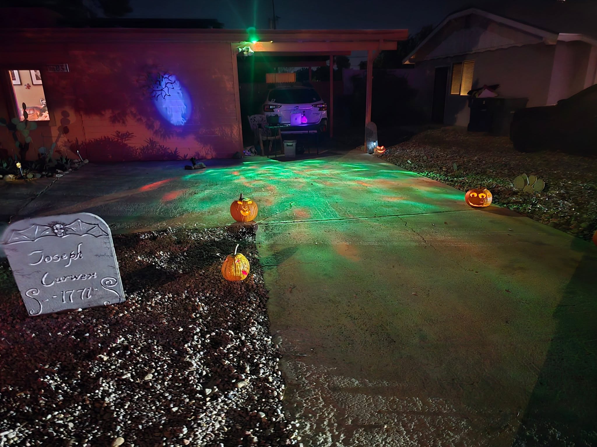A carport set up as a Halloween staging area, with the car pulled deep inside and a lit candy table beside it under the roof.