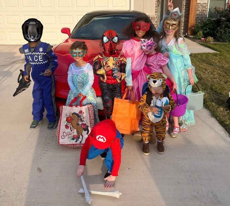 A group of trick-or-treaters of different ages gathered in a driveway at dusk, costumes ranging from a racing suit and superheroes to princesses and a tiger, buckets and tote bags in hand.