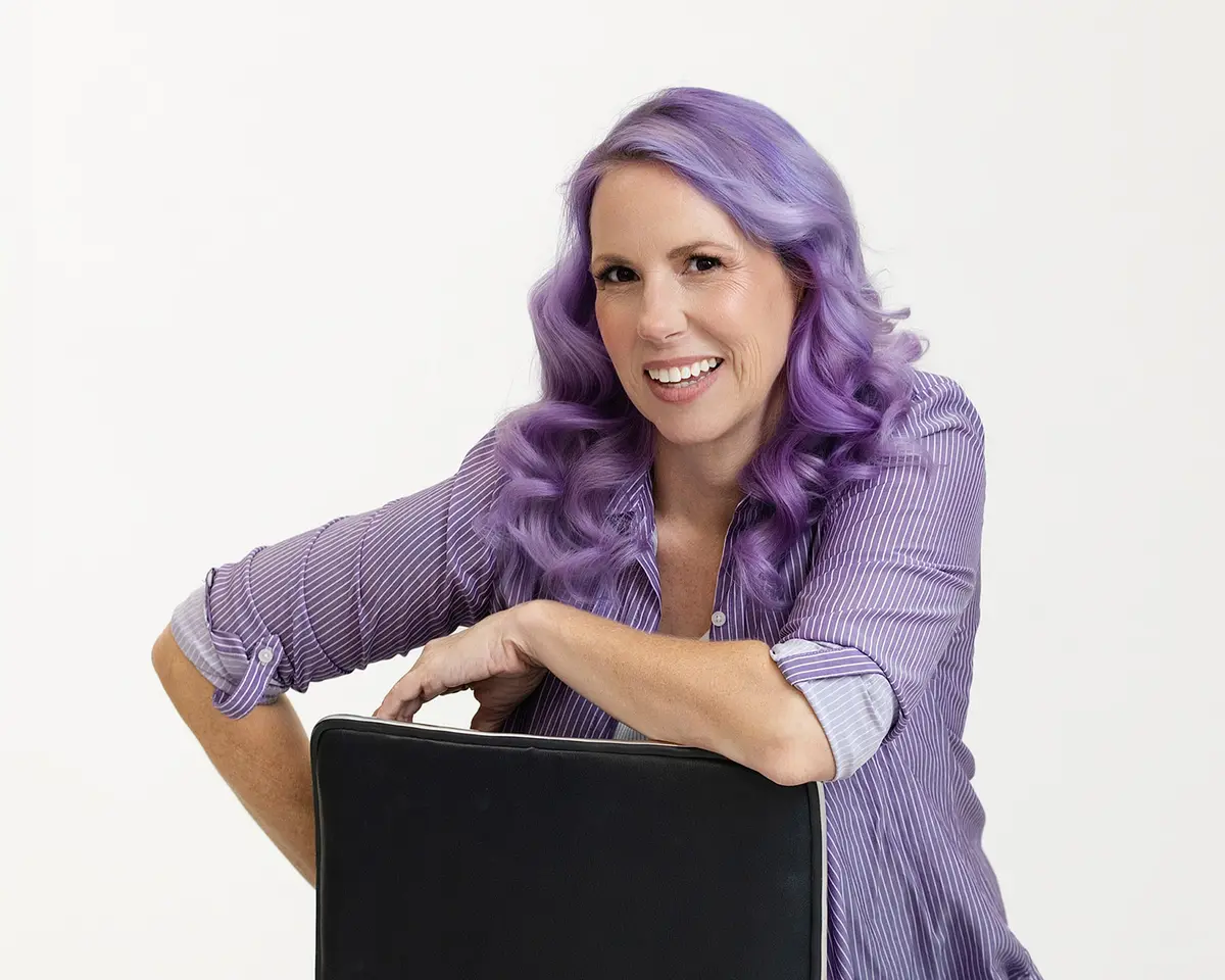 Jennifer Winchester, Tucson real estate agent, smiling and leaning on the back of a chair. She has long wavy purple hair and wears a purple and white striped button-down shirt.