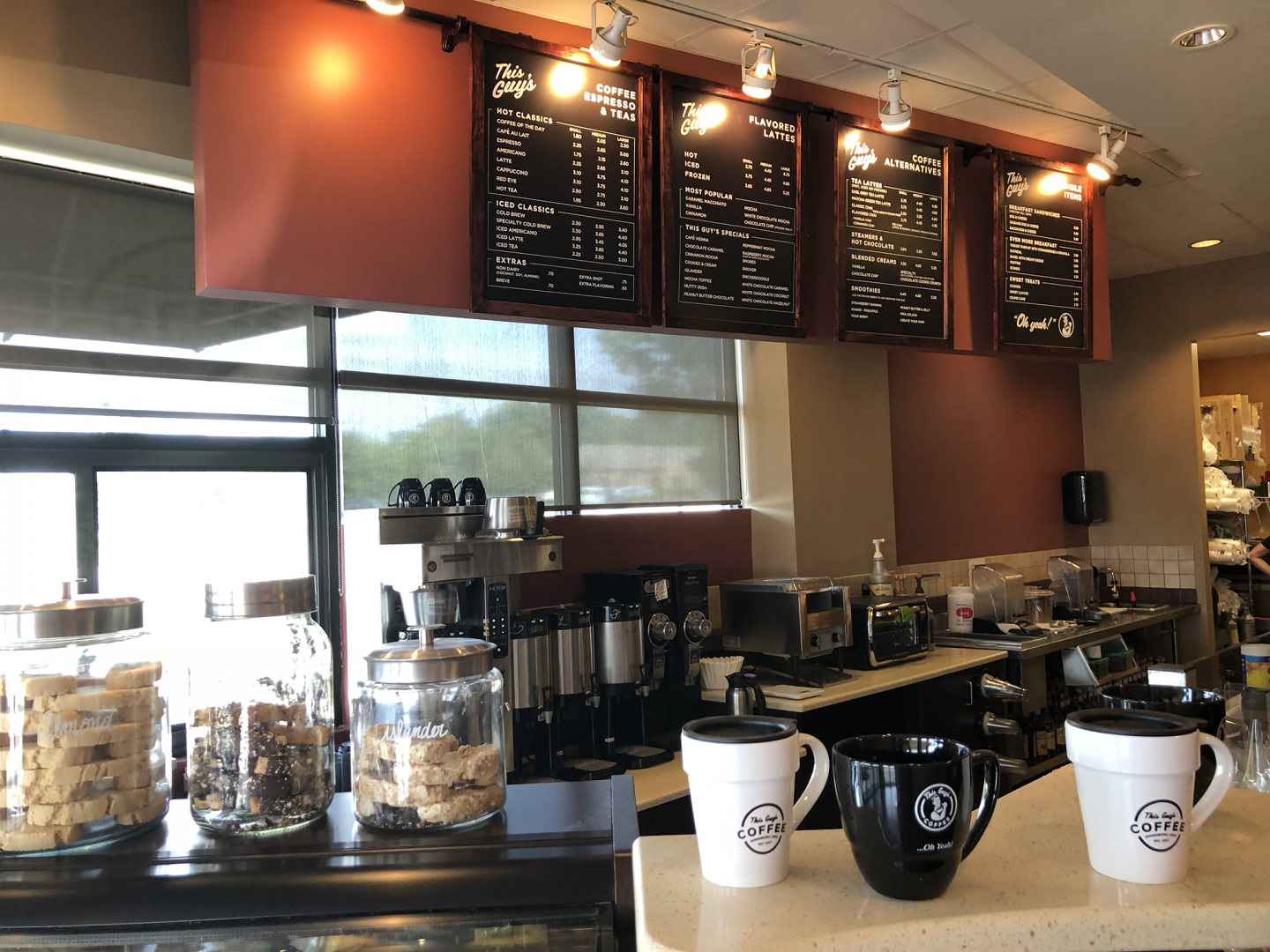 A cozy independent coffee shop interior with a light wood counter, espresso machine, a latte with foam art, a pastry case and hanging plants at This Guy's Coffee in Springboro Ohio
