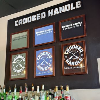 A welcoming craft brewery taproom with a polished wood bar, beer tap handles, exposed brick walls, warm Edison-bulb lighting and copper kettles at Crooked Handle Brewing in Springboro Ohio