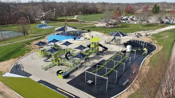 A sunny community park in Springboro Ohio with a wide green lawn, a colorful playground, a curving walking trail past mature trees and soccer fields under a clear blue sky