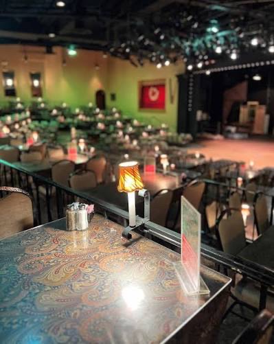 Round tables set with white linens, wine glasses and folded napkins facing a brightly lit Broadway-style stage with red velvet curtains at La Comedia Dinner Theatre in Springboro Ohio