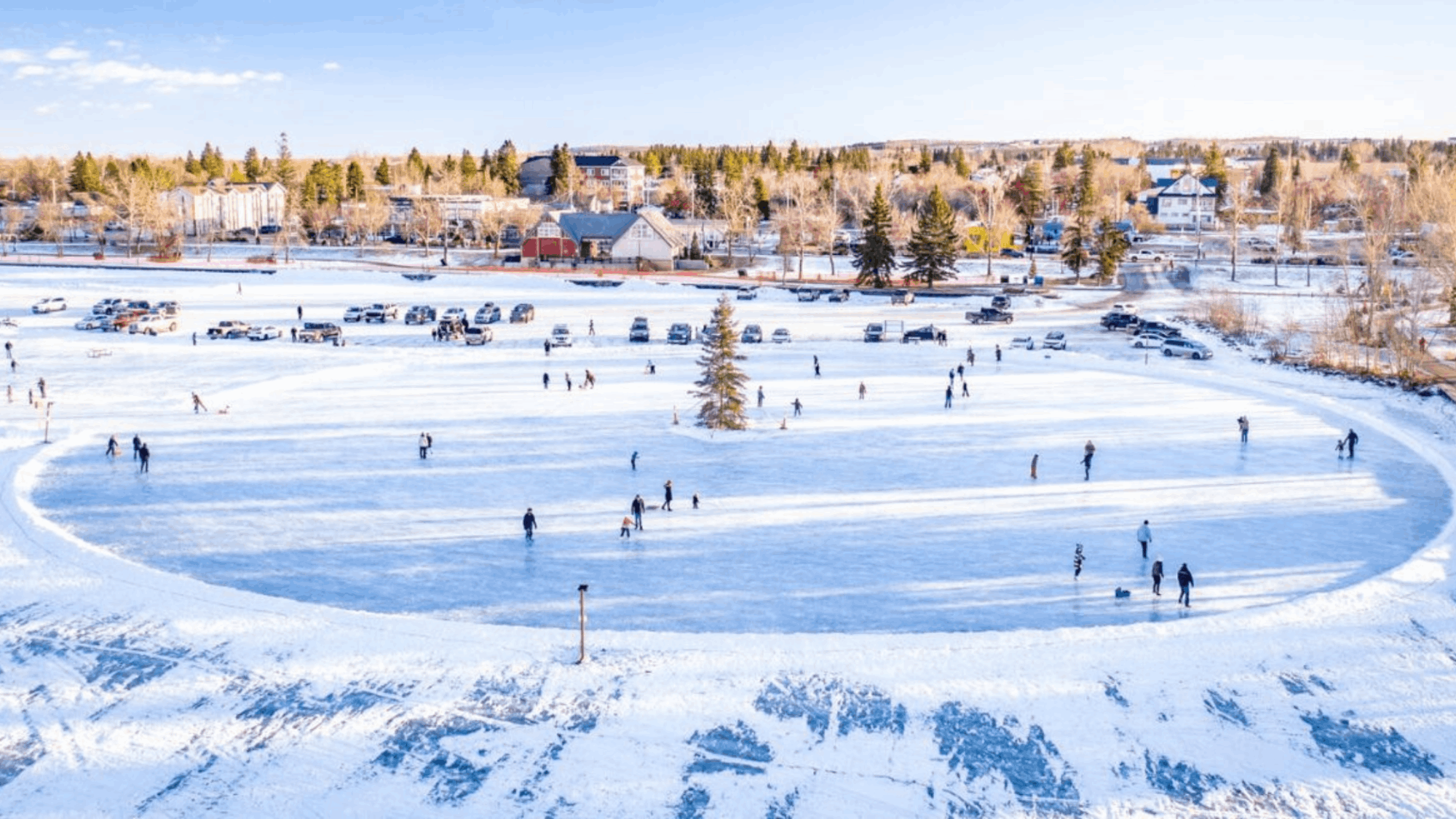 Winter community scene in Central Alberta showing people skating and gathering on a frozen outdoor rink surrounded by neighbourhood homes and parked vehicles.