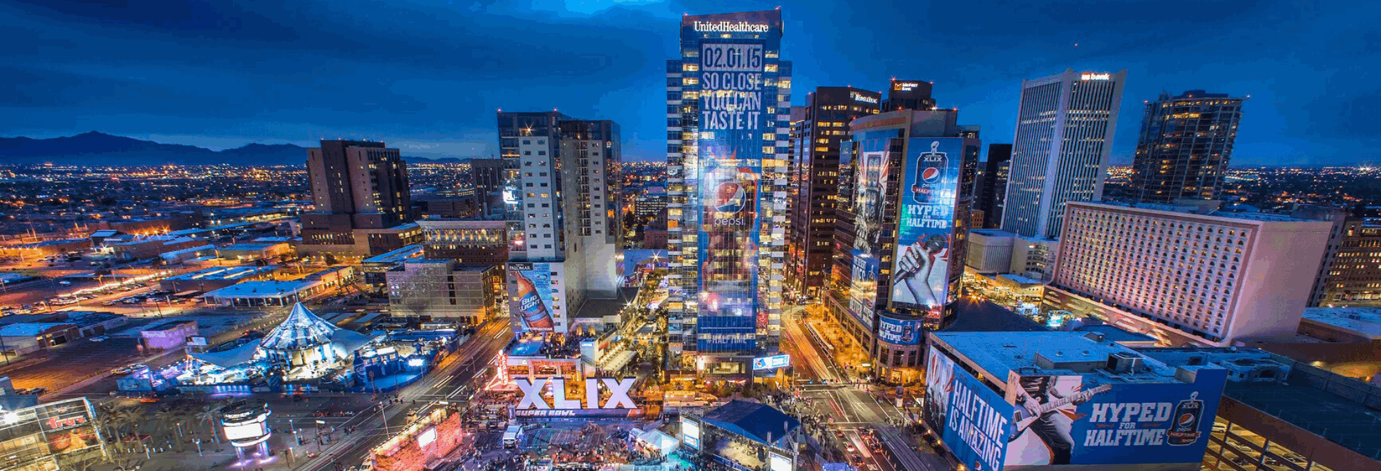 A vibrant aerial view of downtown Phoenix at night, illuminated by colorful city lights and large digital billboards promoting Super Bowl XLIX. The skyline features high-rise buildings, including the UnitedHealthcare Tower with a Pepsi ad, and bustling streets below filled with crowds and entertainment setups. The desert mountains are visible in the background under a twilight sky.