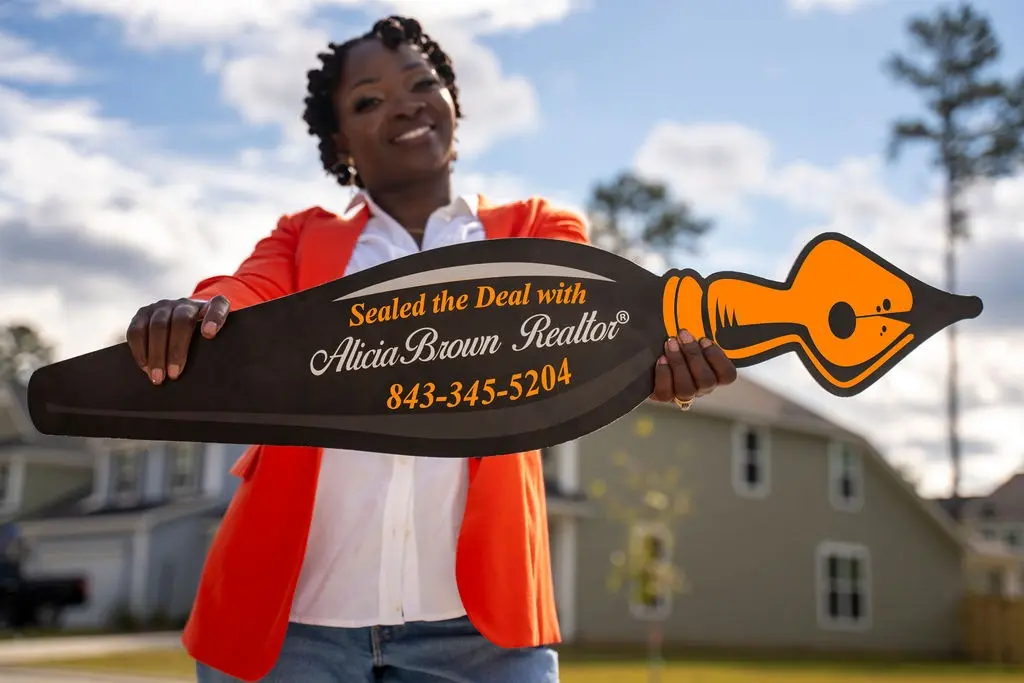 Alicia Brown smiling in a bright orange blazer holds a large, pen-shaped sign outdoors, reading "Sealed the Deal with Alicia Brown Realtor." Suburban homes in the background under a blue sky.
