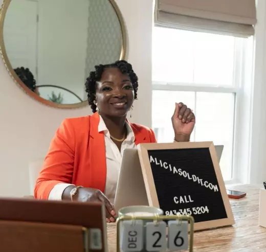 Alicia Brown sitting on a chair with a table smiling.