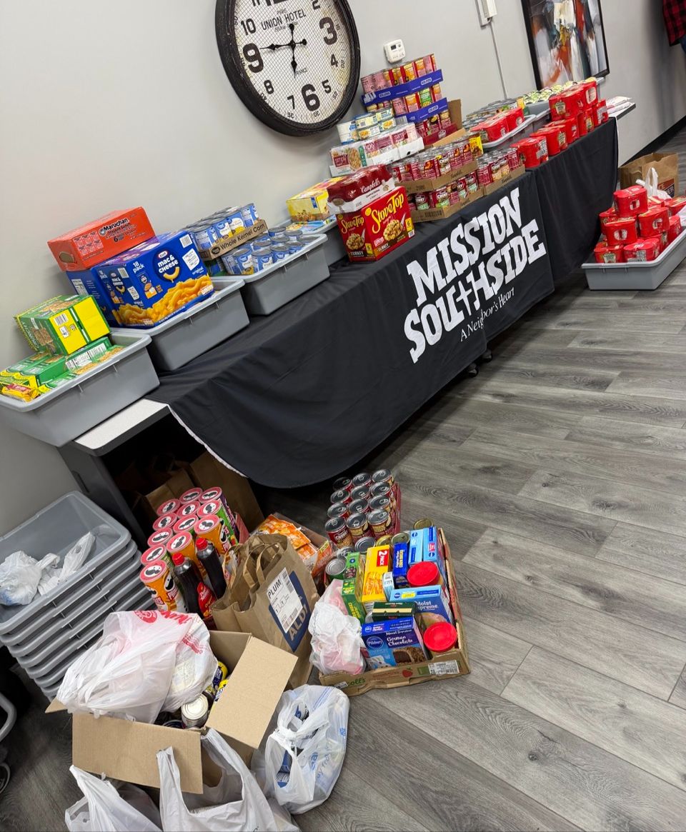 A food donation staging area featuring a table draped with a "Mission Southside" banner, stacked with canned goods and boxed foods, with additional donations piled on the grey wood-look floor.