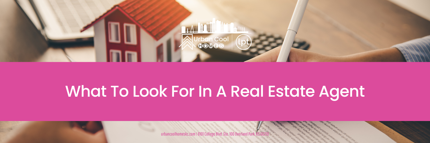 A miniature house model sits on a desk with a laptop, calculator, and a hand holding a pen over a document, with text overlay "What To Look For In A Real Estate Agent."