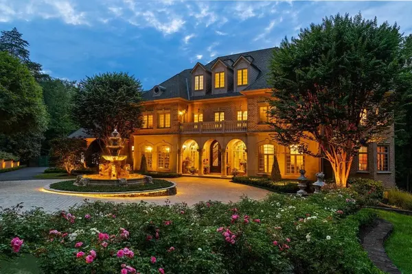 Stately brick manor at dusk with dramatic exterior lighting, arched stone entryway, manicured rose gardens, and illuminated fountain in the motor court