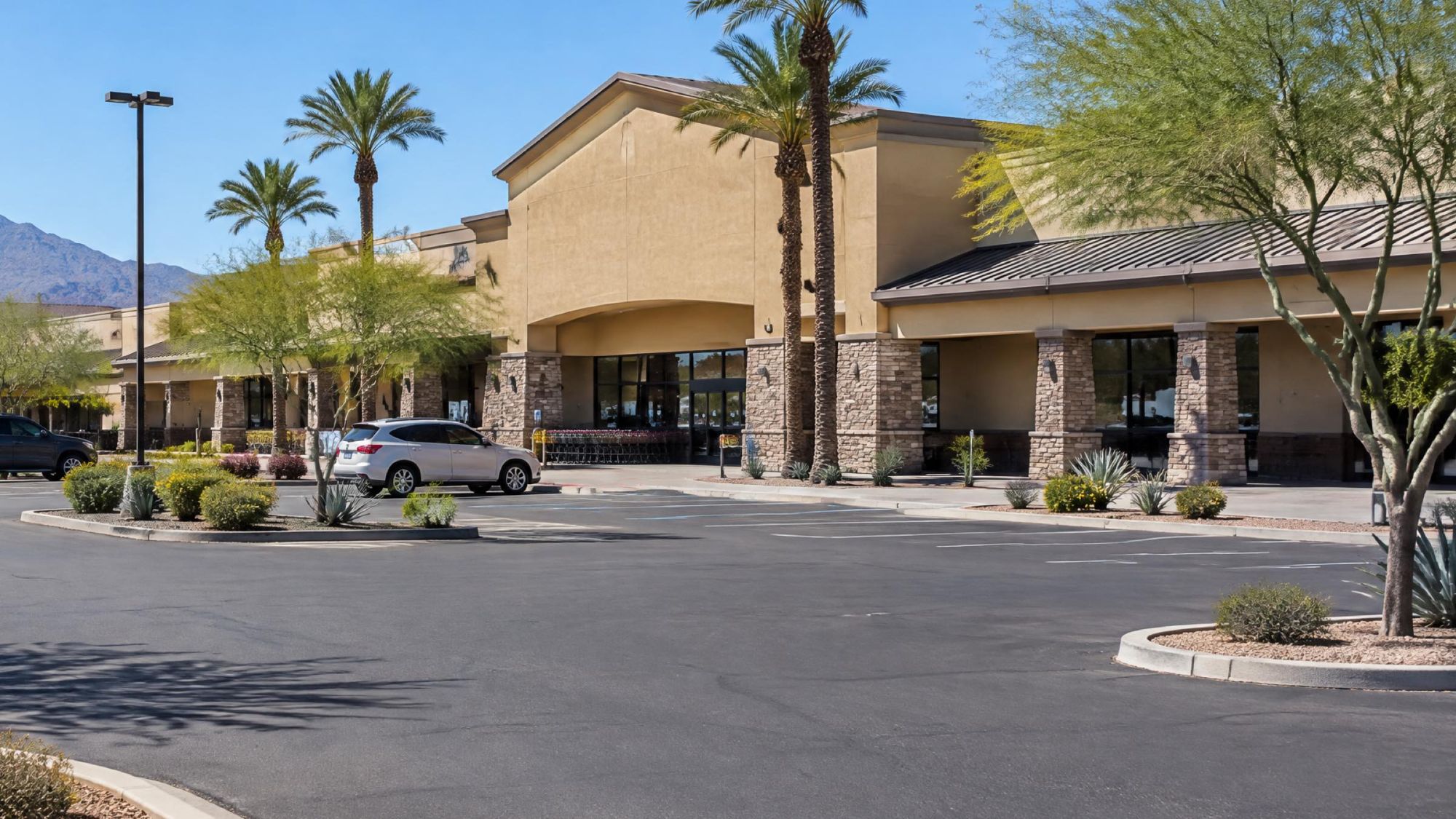 Illustration of a desert shopping center with palm trees and a parking lot, with mountains to the west.
