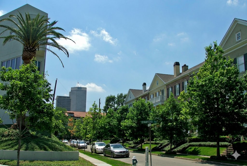 Sunny, tree-lined residential street view in the Montrose neighborhood of Houston, Texas. Features a modern building with a palm tree on the left, traditional brick townhomes on the right, and the downtown Houston skyline visible in the distant background.