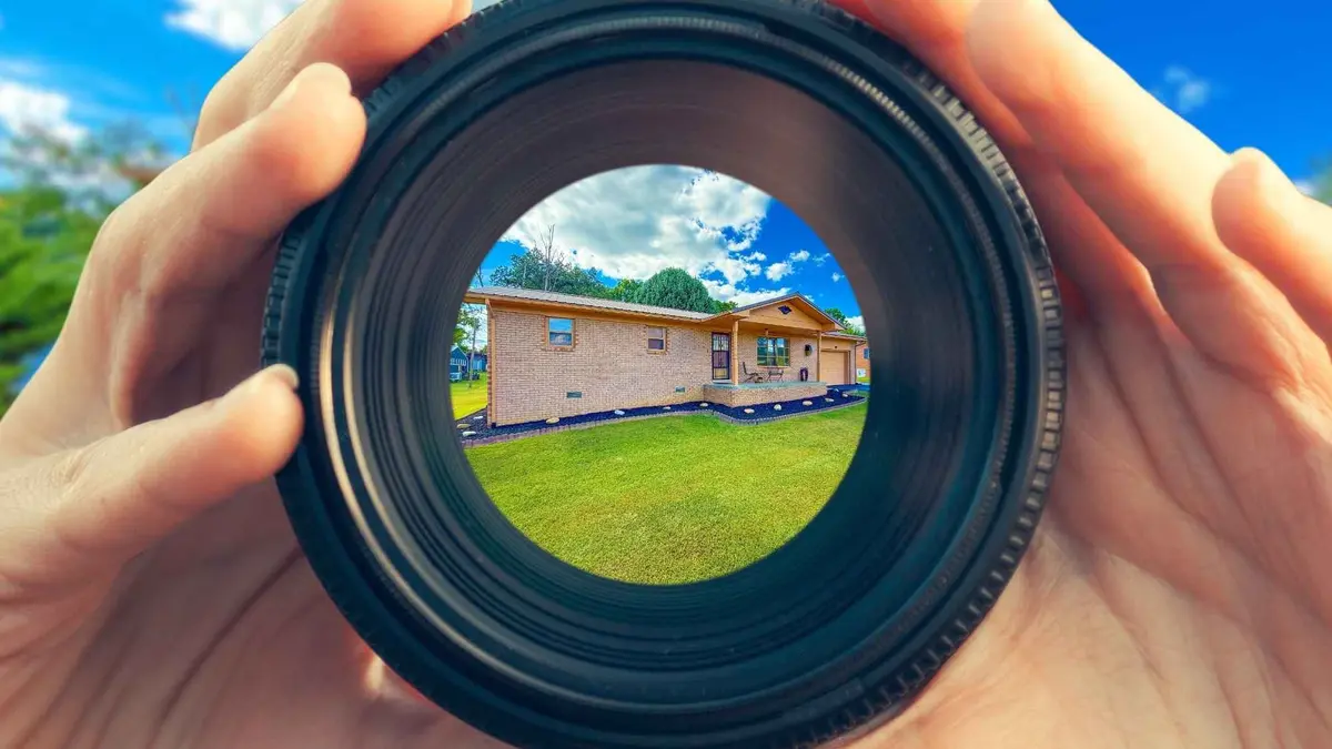 Hands holding a camera lens focused on a brick home with green lawn and blue sky, symbolizing real estate photography and home selling with Williams Elite Realty in Kentucky.