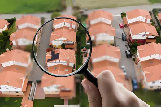 Magnifying glass focusing on a house among several rooftops, representing home search, property listings, and real estate market insights.