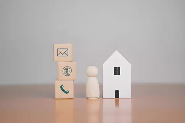 Contact concept showing a small wooden house, a figure, and blocks with email, phone, and message icons — symbolizing real estate communication and customer connection.
