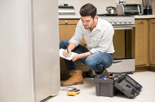 A contractor crouches in a kitchen while writing on a clipboard with tools and a multimeter on the floor