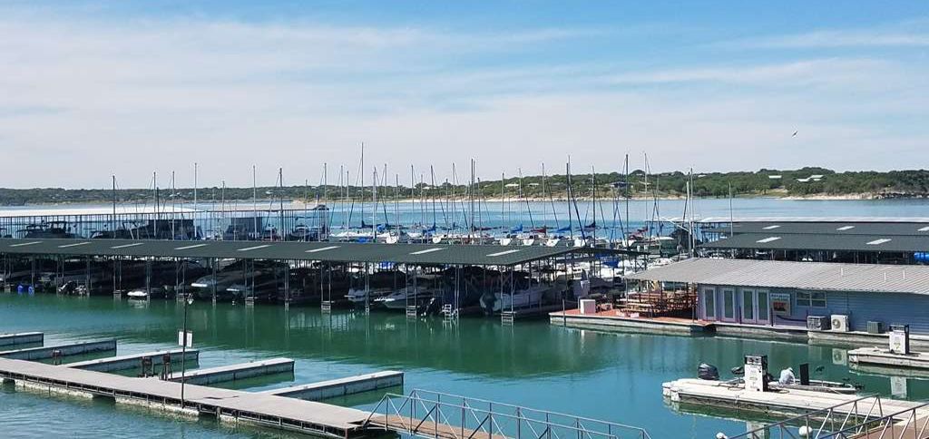 A high-angle view of a Canyon Lake marina featuring covered boat slips, sailboats with tall masts, and the Texas Hill Country shoreline under a clear blue sky.