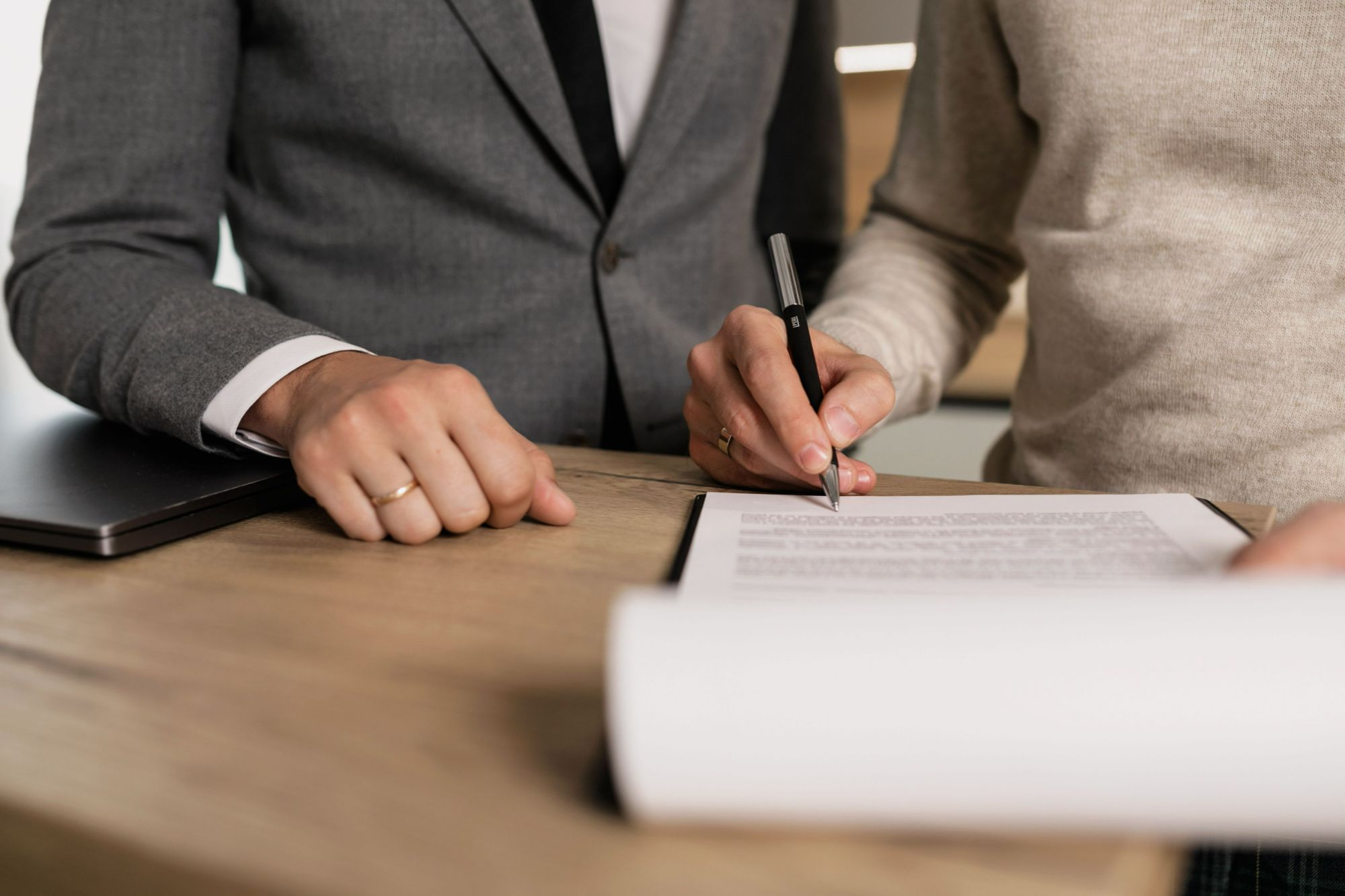 Client signing legal closing documents with a pen while an agent in a grey suit looks on at a wooden desk