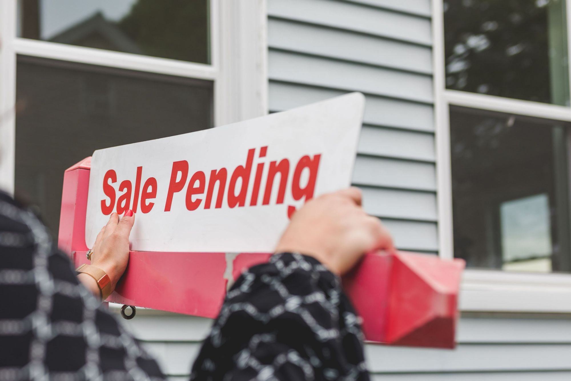 Hands securing a white "Sale Pending" rider sign onto a bright pink real estate post outside a house