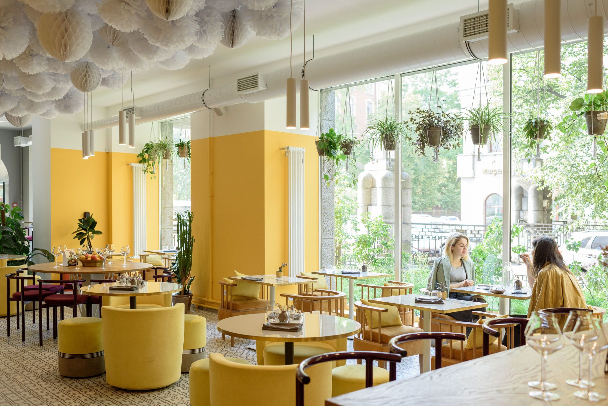 Bright cafe interior with yellow walls, round dining tables, hanging potted plants, and patrons seated near large floor-to-ceiling windows