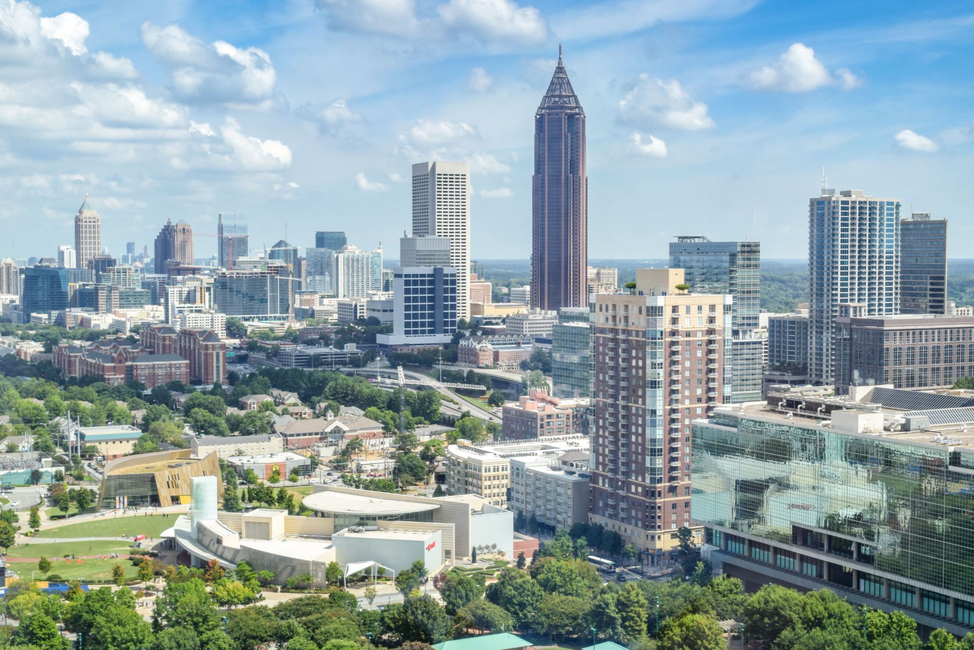 Wide elevated skyline view of Atlanta featuring prominent skyscrapers, green park trees, and mid-rise buildings under a blue sky with white clouds