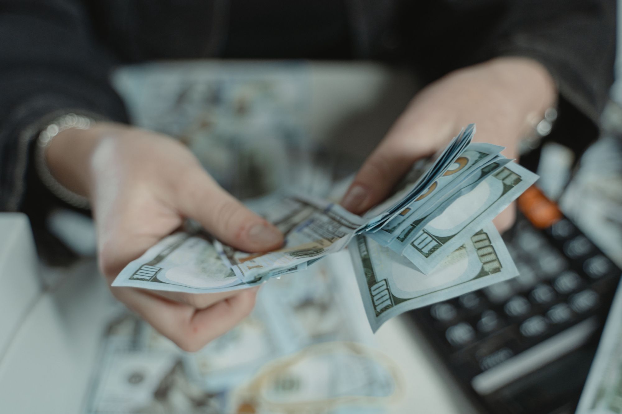 Close-up hands counting a stack of one hundred dollar bills above a desk calculator