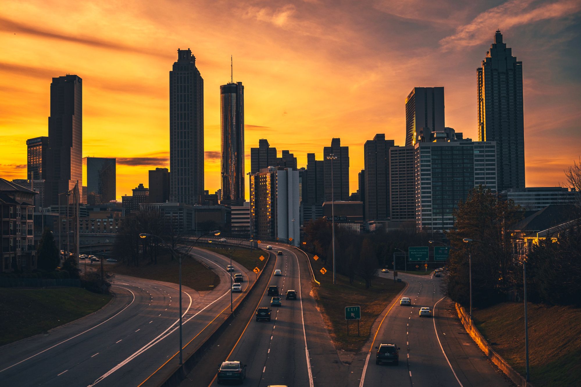 High-angle twilight view of highway traffic curving toward the illuminated Atlanta city skyline beneath an orange sunset sky