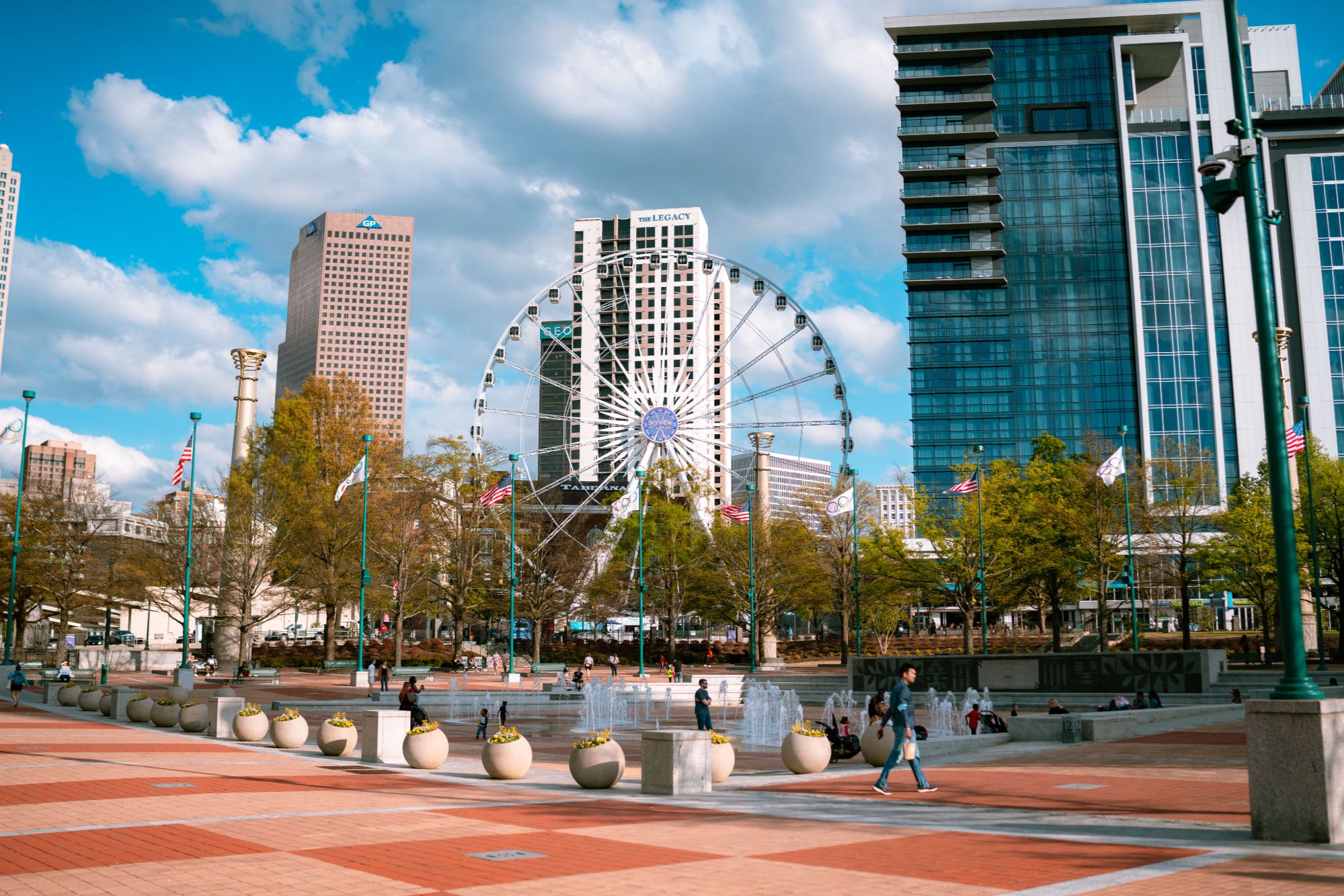 Centennial Olympic Park public plaza featuring water fountains, pedestrians, modern city high-rises, and the SkyView Ferris wheel under a blue sky