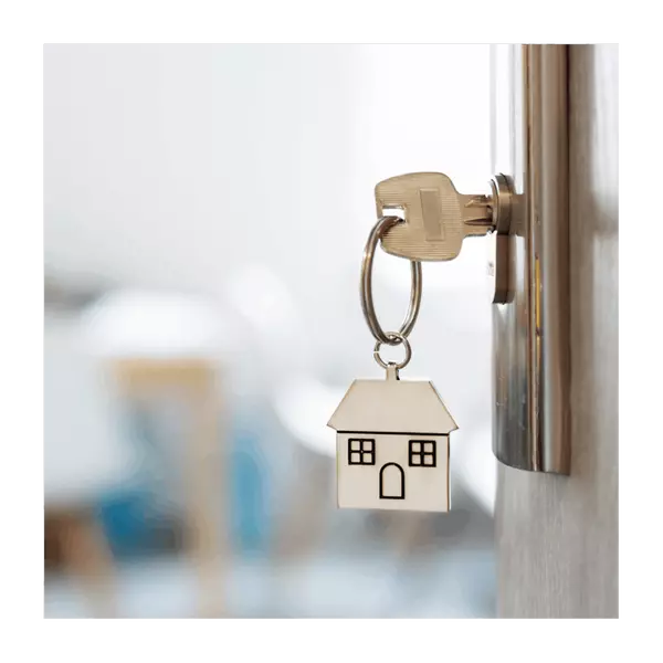 House-shaped keychain dangling from a key inserted into a metal door lock with a blurred interior in the background representing home showings and open houses.