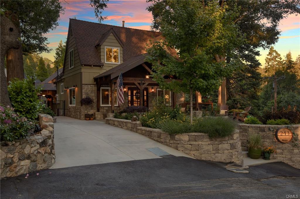 Inviting Lake Arrowhead mountain home at sunset with stone accents, flag, trees, and a long driveway leading to the front porch.