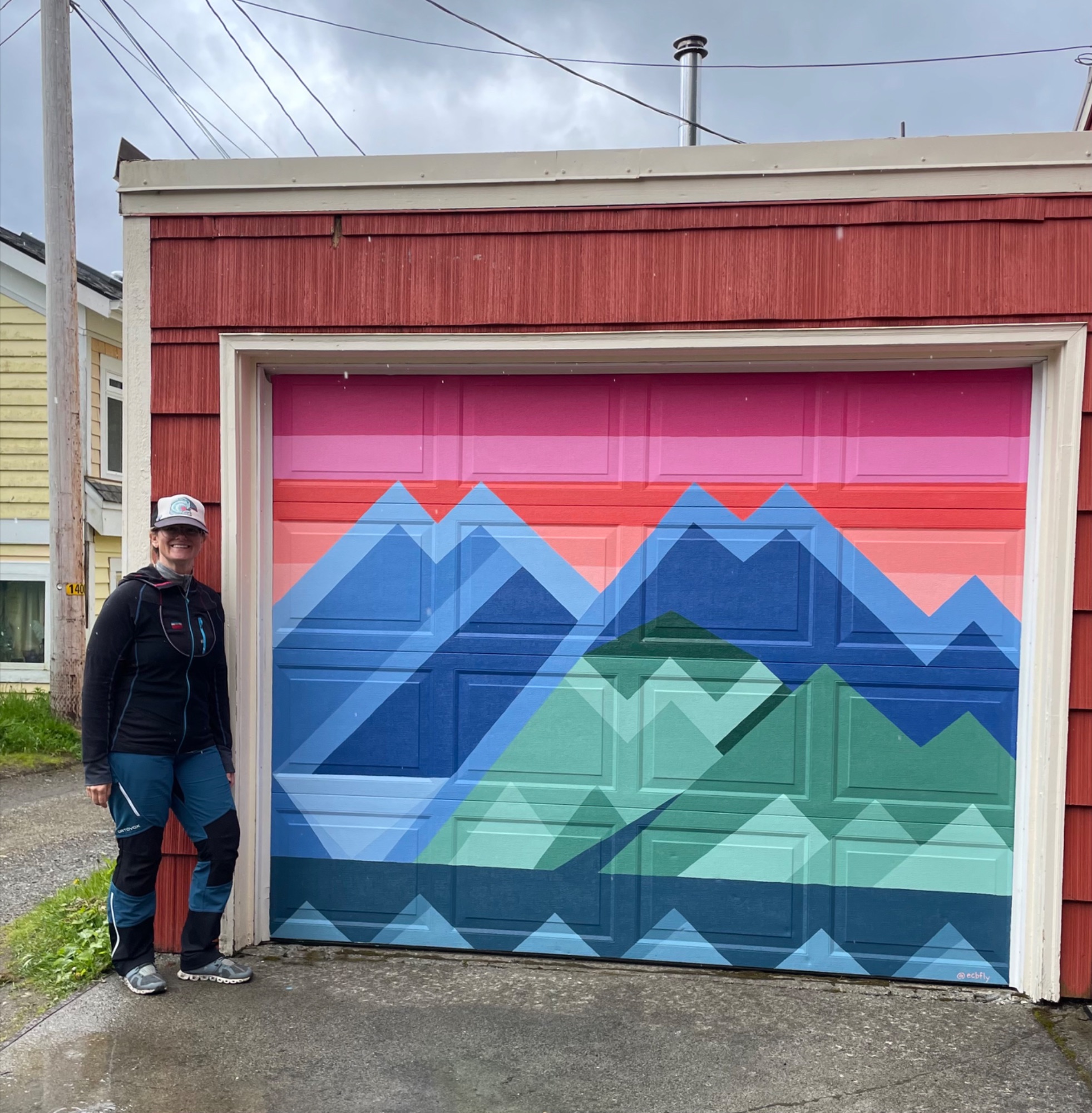 Erin standing next to a geometric mountain mural painted on a garage door with a colorful sunset background.
