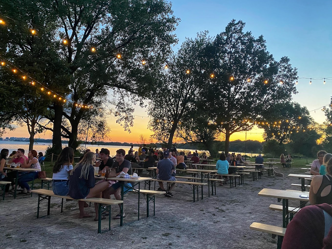 People enjoying food and drinks at the Olbrich Biergarten in Madison, Wisconsin, with picnic tables, string lights, and a sunset view over Lake Monona