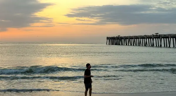 Person walking along the shoreline at sunrise near the Jacksonville Beach Pier, with gentle Atlantic Ocean waves and colorful morning skies in Jacksonville Beach, Florida.