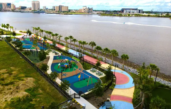 Aerial view of a Jacksonville waterfront park along the St. Johns River with recreation areas, palm trees and the city skyline in the distance.