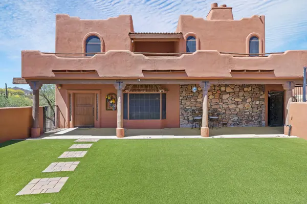 Front view of a private walled courtyard featuring high-quality artificial grass and a southwestern adobe-style home. The exterior includes traditional arched windows, a stone accent wall, and a custom wood entry door.
