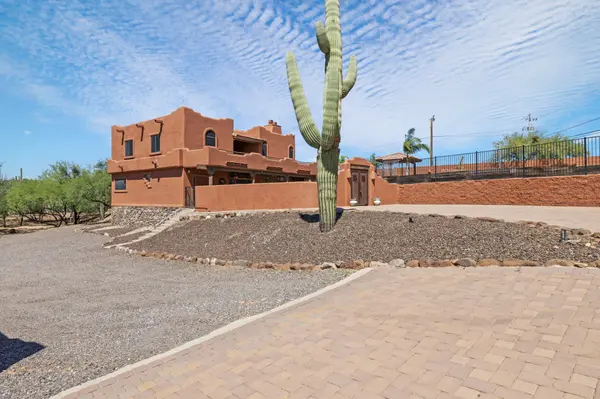 A side exterior view of a multi-level Santa Fe style hacienda in New River, Arizona, featuring traditional wooden vigas and a stone-accented foundation. The desert landscape is meticulously maintained with crushed rock and a large Saguaro cactus, framed by a custom paver driveway and a red block retaining wall under a bright, wispy blue sky.