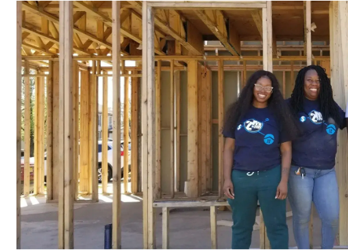 Two women standing inside wooden frame construction at a building site, portrait-oriented photograph showing framing and partial exterior lighting, approximate aspect ratio 1:1