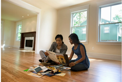 Two women sitting on a hardwood floor in an empty house looking through papers and magazines, bright window light and fireplace visible, approximate aspect ratio 7:6