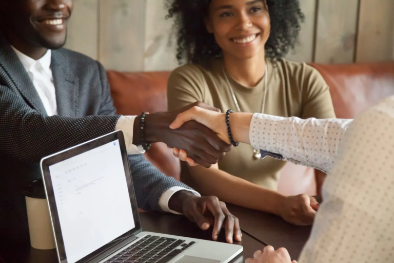 A small group seated around a laptop, hands extended in a handshake-like gesture, suggesting a planning meeting in a casual office setting.